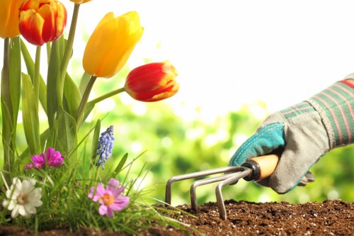 A close-up view of a gardening scene in a residential outdoor space, featuring a person wearing a gardening glove using a hand fork tool to cultivate dark, fertile soil near blooming flowers. The flower bed includes vibrant tulips in shades of red and yellow, along with smaller pink, white, and purple flowers, set against a background of lush green foliage and a bright, sunlit sky. The garden area is part of a landscaped yard typical of Leyton, with a well-maintained lawn and clean paving visible in the background. The scene emphasizes careful planting and soil preparation, relevant to gardening services such as lawn and flower bed maintenance offered by Lawn Mowing Leyton, situated in the Leyton postcode area, promoting outdoor garden care in east London.