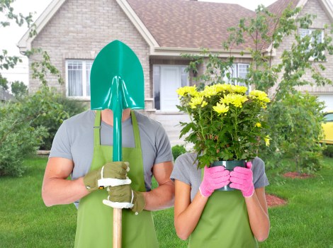 A gardening scene in a residential garden during daytime shows two people wearing green gardening aprons and gloves. One person, dressed in a light grey shirt, holds a blue and green garden shovel with the blade facing upwards, partially obscuring their face with a green gardening hood. The other person, in a grey shirt with pink gloves, holds a large pink pot filled with vibrant yellow chrysanthemums, which cover most of their face. The garden features a well-maintained lawn with lush green grass, bordered by shrubs and small trees. In the background, there is a brick house with white-framed windows, a small front porch, and a paved pathway leading to the entrance. The environment looks bright and clear, suggesting a calm, sunny day suitable for outdoor gardening work, and the scene exemplifies landscaping and garden maintenance activities typical of Leyton, supporting local garden care services.