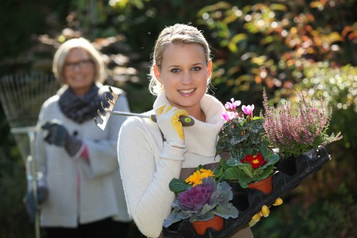 A young woman with blonde hair tied back, dressed in a cream sweater and gardening gloves, is standing outdoors in a lush garden holding a black tray filled with colorful flowering plants and green foliage. The garden features a well-maintained lawn with dense trees and shrubs in the background, displaying various shades of green and hints of autumnal colours. Behind her, an older woman with short grey hair, glasses, a white coat, and a dark scarf is holding a garden tool, smiling as she observes the scene. The setting appears to be in a private garden or landscaped outdoor space, with sunlight filtering through the trees, creating a warm and inviting atmosphere. The garden elements, including the vibrant flowers, neatly trimmed plants, grass, and surrounding greenery, reflect professional gardening and landscaping care, which Lawn Mowing Leyton offers in Leyton, addressing lawn maintenance and garden enhancement needs.