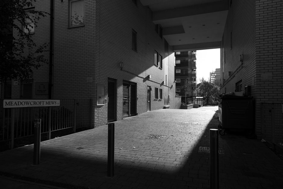 A narrow alleyway between two modern brick apartment buildings, with a paved surface and a slight incline. The alley is partially shaded by the overhanging structures of the buildings, creating a brig